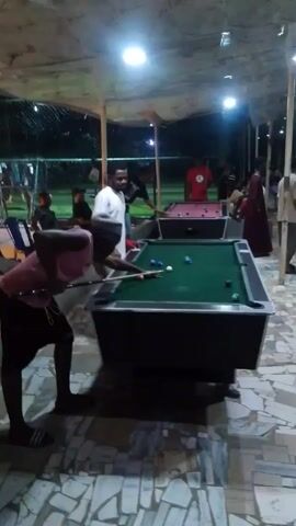 The video shows a group of people under a roofed patio structure at night, playing pool. A person in a pink shirt and shorts, wearing a grey head covering, is leaning over a pool table, aiming with a pool cue at the cue ball. There are multiple colored pool balls on the table. Other people are standing around or seated in chairs. Another pool table is visible in the background, where more people are playing. The patio floor is tiled with light-colored stone. Lighting comes from overhead ceiling lights and from outside the patio. There is a fence visible outside. The overall impression is of a casual, social gathering.
