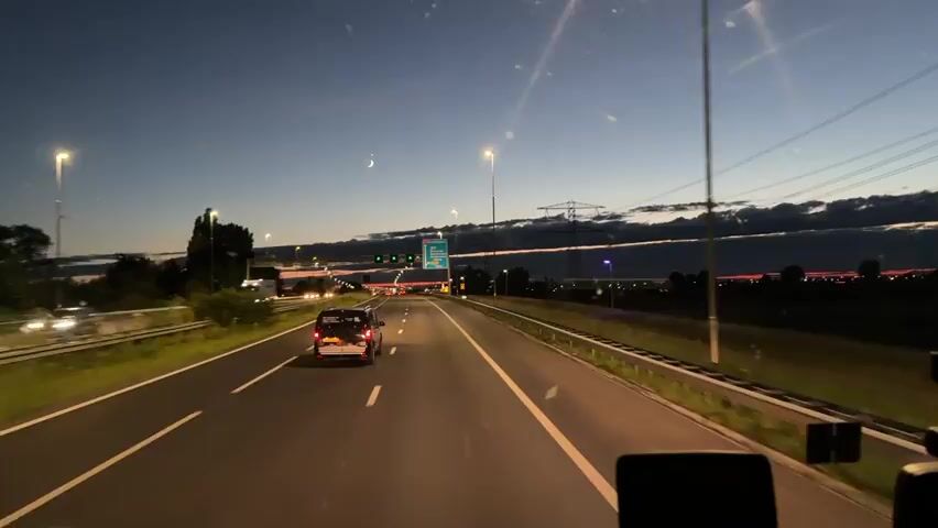 A first-person perspective video shows driving on a highway at dusk. The sky is a deep blue with some clouds and a crescent moon. There are streetlights and traffic lights visible, as well as power lines and towers. Several cars are on the road, including a truck in the left lane. The camera is mounted inside a vehicle, possibly a truck or bus, with a dashboard visible at the bottom of the frame. A signpost with Dutch text and arrows indicates directions to various towns. The video captures the transition from day to night on a busy highway.