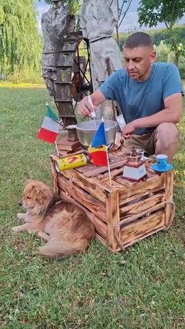 A man is kneeling in a grassy area outdoors, stirring a pot with a whisk. In front of him is a wooden crate with a yellow can, two small flags (one Italian, one Romanian), a coffee grinder, and a blue teacup. A light brown dog is lying on the grass to the left of the crate. Behind the man and crate is a large stone sculpture or water feature. The man is wearing a blue t-shirt. The scene appears to be a rustic setting.