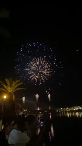 This video captures a nighttime fireworks display over a body of water, likely the Gilão River in Tavira, Portugal. The scene is illuminated by the vibrant bursts of fireworks, predominantly in shades of gold and white, with some green accents. The dark sky serves as a dramatic backdrop for the exploding pyrotechnics.

In the foreground, silhouetted figures of people are visible, gathered to watch the spectacle. Some are holding up their phones, presumably to record the event. The presence of a palm tree on the left side of the frame and the distant lights of buildings along the water's edge provide clues to the location. The reflection of the fireworks and lights on the water creates a shimmering effect, adding to the festive atmosphere.

The time of day is clearly night, and the event taking place is a fireworks show, likely a celebration or festival. The overall mood appears to be one of awe and enjoyment, with the bright lights and gathering of people suggesting a communal experience. No visible text is present in the video.