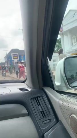 The video is shot from the interior of a vehicle, looking out through the front passenger side window. The vehicle's dashboard, a vertical air vent, the A-pillar, and a portion of the door panel with a circular speaker grill are visible in the foreground. Outside the vehicle, an urban street scene unfolds under an overcast sky. On the right side, a white building with pillars is partially visible. Further down the street on the left, a blue building with a purple "FCMB" sign is present. Multiple pedestrians are walking and standing on the street and sidewalks. Several motorcycles are visible, some appearing stationary. The vehicle's side mirror reflects elements of the outdoor environment. Voices are audible, with one person mentioning "FCMB" at 0:04.