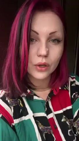 A woman with bright pink and black hair wearing a patterned shirt is smoking a cigarette. She is indoors, facing the camera directly.  She brings the cigarette to her lips, inhales, and exhales a cloud of smoke. Her left hand, which holds the cigarette, has multiple tattoos.