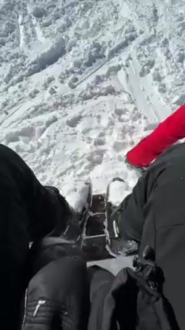 Here is a description of the video.

The video shows a person sitting in ski boots on a snowy surface. A large sign reading "La Clusaz" is visible in the background. The camera pans up to show a person in a red ski suit standing near the sign. They have on a helmet with bear ears and appear to be looking at the view. The sky is blue with some clouds. Trees covered in snow are also visible.
