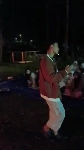 At night, a person stands in a dimly lit outdoor area, facing a group of seated individuals. The person is wearing a brown and white jacket, light pants, and dark shoes. They are gesturing with their hands as if speaking or giving a presentation.  The seated individuals appear to be mostly female, some wearing hijabs. A fire burns brightly in the foreground, providing some illumination to the area.  In the background, there are trees, a building with lights, and a few other figures. The ground appears to be covered in grass or dirt.