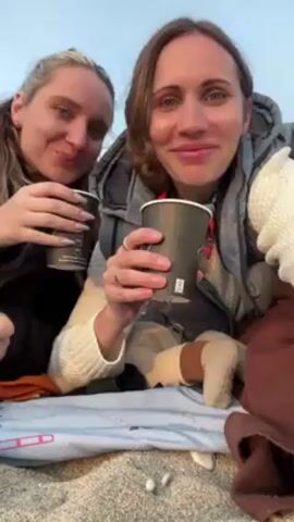 Two women are sitting on a blanket outdoors. They are both holding black paper cups and smile at the camera before taking a drink. The woman on the left has blonde hair and a black jacket. The woman on the right has brown hair and wears a gray vest. Both women have white sweaters underneath their outer layers, and they both wear rings on their left hands. A child's legs, with socks, are visible between them.  A brown jacket is draped over part of the blanket. They are on a beach with a light blue sky visible in the background.
