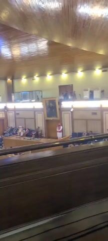The video shows the interior of a parliamentary hall with wooden panelling. There are rows of seats facing a central platform where a man is addressing the assembly. A large screen displays the speaker's image. Several men are seated in the hall, listening to the speech. One man in a blue denim jacket walks past the camera. The scene suggests a parliamentary session or a formal meeting.