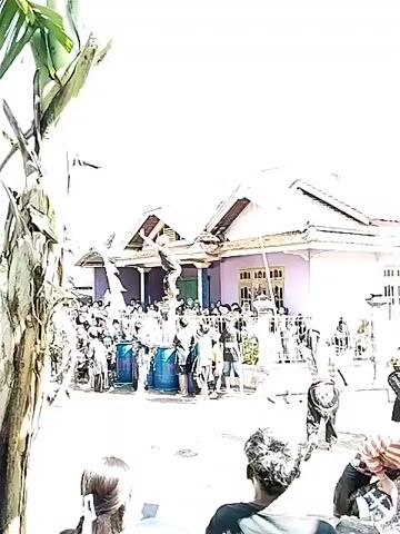 A crowd of people has gathered in front of a purple house with a brown roof to watch a performance. A person dressed in black and red stands on top of several blue barrels, making acrobatic movements. There is a tall bamboo pole in the foreground. People in the crowd are taking pictures with their phones. The weather is sunny. A banana tree stands to the left.