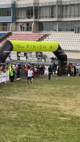 The video captures the start of a marathon or race under an inflatable archway that reads "ALMATY MARATHON FINISH". The arch is black and lime green, with sponsor logos including "SALOMON", "345", and "ASIAN MOTORS". A crowd of participants, mostly children and young adults, are gathered behind a barrier, about to begin running. Spectators are visible on the sidelines, some cheering and holding up phones. The event appears to be taking place in an outdoor stadium with tiered seating in the background. The grass is green and well-maintained. The sky is overcast, suggesting a cloudy day. The overall mood is energetic and anticipatory as the runners prepare to start their race. The text "TASS" is also visible on the archway, likely indicating a media partner. The setting suggests a public event in a sports venue.