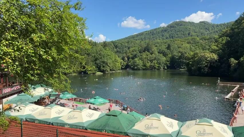 The video opens on a wide shot of a natural lake surrounded by lush green trees and a distant mountain under a bright blue sky with scattered clouds. In the foreground, rows of green and white umbrellas are set up over wooden decks and tables, some with lounge chairs. People are visible relaxing under the umbrellas and on the decks. The lake itself is populated with numerous individuals swimming and floating. Some are gathered near a wooden platform extending into the water, while others are scattered throughout the lake. A building, possibly a lodge or changing facility, is situated on the right side of the lake, with steps leading down to the water. The scene suggests a popular recreational area on a sunny day. As the video progresses, the camera pans slightly to the right, offering a clearer view of the building and more activity on the decks.