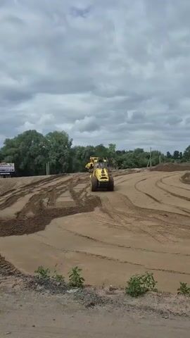 Two yellow construction vehicles are operating on a dirt surface. A bulldozer is in the background, and a smaller, wheeled vehicle, possibly a front-end loader, is in the foreground. The wheeled vehicle is moving and leaving tracks in the dirt. The ground appears to be a mix of sand and dirt, with some loose piles. Trees and vegetation are visible in the background, and the sky is overcast with clouds. The overall impression is a construction site in progress, likely involving earthmoving or grading.