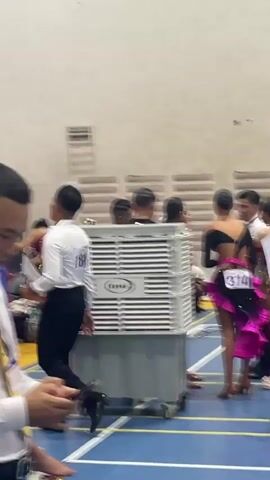A group of people are in a brightly lit indoor space that appears to be a gymnasium or dance studio. The floor is a blue surface with yellow lines. In the foreground, a large white industrial-looking air cooler with the logo "DHF" is visible. Several individuals, mostly young women in colorful, sequined dance attire, are present. Some are standing and talking, while others appear to be preparing or waiting for an event. One woman in a red sequined dress with a high slit is walking towards the right side of the frame. Another woman in a similar red dress is visible further back, also in motion. A few people in casual clothing, including a person in a grey top and pants and a person in a black t-shirt, are also scattered among the dancers. One person is crouched down near the air cooler. In the background, there are walls with horizontal slatted elements and a sign that reads "WC". The overall impression is of a backstage or waiting area for a dance competition or performance.