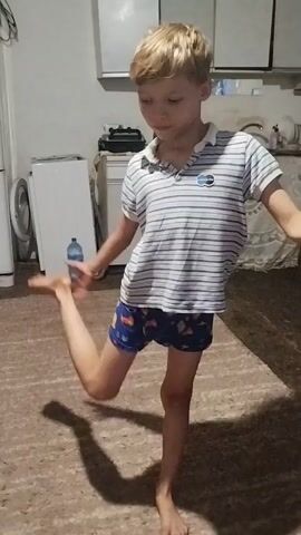 A young boy with blond hair is standing on one leg with the other leg bent and his foot held by his hand in what appears to be a kitchen. He is wearing a striped polo shirt and patterned shorts. The floor is carpeted and there is a washing machine to the left.