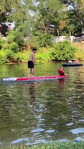 A man and a child are on a paddleboard on a body of water. The man is standing and paddling with a green paddle. The child is sitting on the back of the paddleboard wearing a life jacket. There are other people on the water in inflatable tubes in the background. The environment is a river or lake with trees and foliage on the shore.