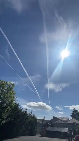 The video shows a clear blue sky with the sun shining brightly, surrounded by some light clouds. Several white contrails from airplanes streak across the sky, intersecting at one point.  Below, the tops of trees and roofs of buildings are visible, indicating an urban or suburban environment. A red car is visible in the lower right corner of the frame.