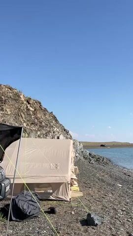 The video shows a tent set up on a rocky shore next to a body of water. The sky is clear and blue. In the background, a large rock formation rises steeply. A bird of prey is perched on the top of the rock formation. The camera zooms in on the bird. The person filming asks if the bird is visible through the fog, to which another person replies that it is not.