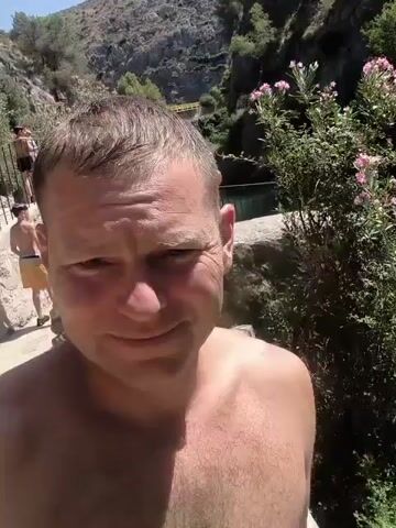 The video opens with a close-up of a man's face as he smiles into the camera. He is shirtless and appears to be outdoors. The background shows a natural landscape with rocks and vegetation. As the video progresses, the camera pans to reveal a waterfall and a body of water below. There are other people visible in the background, some standing near the edge of the waterfall and others on the rocky cliffs. The overall scene suggests a recreational setting in a natural environment.