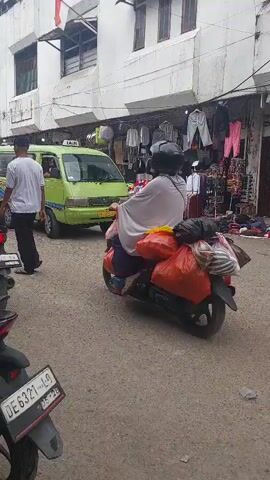 The video shows a busy street scene in what appears to be a marketplace. In the foreground, a person wearing a white t-shirt and grey shorts is sitting on a black motorcycle, facing away from the camera. Another person wearing a red helmet and an orange jacket is standing next to them, also on a motorcycle. Behind them, a green van is visible. The street is lined with stalls selling clothing and other goods. People are walking around, and several other motorcycles are parked or moving. The overall atmosphere is one of commercial activity and everyday life in a crowded urban environment. The ground is paved, and the buildings are multi-story structures with white facades. A red and white flag is visible hanging from one of the buildings. The lighting suggests it is daytime.