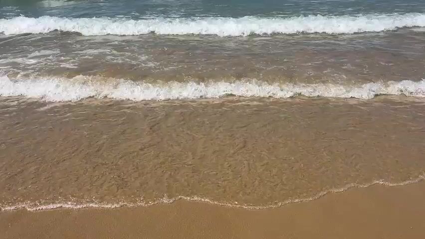 The video shows a close-up, eye-level view of gentle ocean waves rolling onto a sandy beach. The water is clear and shallow, revealing the sand beneath. The waves are small and foamy, creating a peaceful and serene atmosphere. The scene is a typical beach environment, with no people or man-made objects visible. The focus is solely on the natural movement of the water and its interaction with the sand.