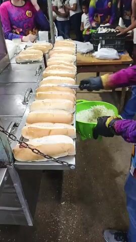 A person in jeans and a purple shirt is preparing hotdogs in an outdoor setting. The person is wearing black gloves and using tongs to put cabbage from a green bucket onto the open hotdog buns, which are lined up on a stainless-steel table. Another person in a purple shirt can be seen behind the table, also working on the hotdogs. Several people are standing around watching or waiting. There is a wooden table behind the stainless-steel one with a black crate on it. The ground appears to be slightly damp with wood shavings scattered about. The scene is well-lit, but there are shadows around the edges, suggesting it is nighttime. Loud music is playing in the background.