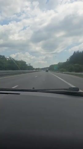 The video shows a first-person view from inside a car driving on a highway. The sky is cloudy with some patches of blue. Green trees line the sides of the highway. Several cars are visible on the road. A white SUV passes on the left lane. A green sign is visible on the right side of the highway. The dashboard of the car is in the foreground.