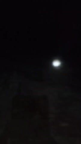 The video shows a dark, blurry nighttime scene. A faint, fuzzy light source is visible in the upper right portion of the frame. The rest of the frame is predominantly black with some indistinct shapes that may be ground or objects. The image is grainy and out of focus, making it difficult to discern specific details. The light source appears to be moving slightly or the camera is unsteady, causing a subtle blur. There are no discernible people, animals, or structures in the video. The overall impression is of a dimly lit, possibly outdoor environment at night.