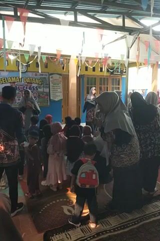 The video shows a group of children and adults, some of whom appear to be teachers, in what looks like a classroom or a community center. Many of the children are wearing hijabs, suggesting a religious or cultural context. The room is decorated with colorful banners and flags. The people are mostly kneeling on the floor, some facing a teacher who is also kneeling. The overall atmosphere appears to be one of a structured activity, possibly a lesson or prayer session. The lighting suggests it is daytime, with sunlight streaming in from an open area. There is a sign on the wall that says "SELAMAT DATANG TRIAL CLASS." The video is filmed vertically and is shaky, indicating it was likely recorded on a mobile phone.