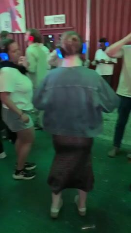 The video shows a group of people at an outdoor event, possibly a party or festival. They are all wearing headphones that light up with different colors, indicating a silent disco or a similar event. The background features wooden walls and large posters, one of which displays the Norwegian flag and a graphic of a person. The ground is illuminated with green and red lights, suggesting a festive atmosphere. People are standing, talking, and some appear to be dancing. The lighting is low, and the overall impression is one of a lively, communal gathering.