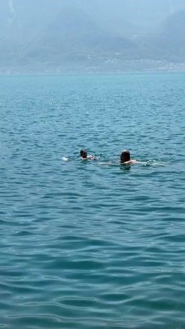 This is a vertical, medium-shot video of two people swimming in a large body of water. The water is a deep blue and appears calm with gentle ripples. In the background, a mountain range with lush green slopes rises majestically. The sky above is a clear, bright blue, suggesting a sunny day.

The two individuals, an adult and a child, are swimming side-by-side. They are both partially submerged in the water, with their heads and shoulders visible. The adult, on the right, appears to be looking towards the child. The child, on the left, seems to be looking ahead. Their body language suggests a relaxed and enjoyable activity, likely a casual swim.

The scene is set in a picturesque natural environment, with the expansive water and imposing mountains creating a sense of tranquility and vastness. The clear sky and bright light indicate it is likely daytime, possibly mid-morning or afternoon. The presence of a child swimming with an adult implies a recreational or family outing.

The visual cues, such as the style of the mountains and the clear blue water, are consistent with a location like Vevey, Switzerland, which is known for its beautiful lake and surrounding alpine scenery. There is no visible text in the video. The overall emotion conveyed is one of peace, joy, and the simple pleasure of being outdoors in a beautiful natural setting.