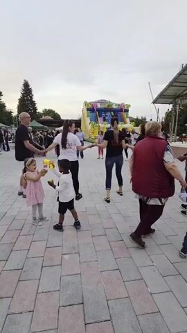 Here's a description of the video:

The video captures a group of people dancing in a circle on a brick-paved area, likely at an outdoor event or festival. There are about 10 adults holding hands and moving to music, along with a few children trying to follow the dance.

In the background, there is a large, colorful inflatable bounce house, indicating a family-friendly event. To the right of the dancers, there is a stage with a partial roof structure and decorations. There are other people milling around the area, suggesting a larger gathering.

The sky appears overcast, and there are trees and other structures visible in the periphery. The overall impression is a relaxed, communal atmosphere with people enjoying traditional dancing at an outdoor celebration.