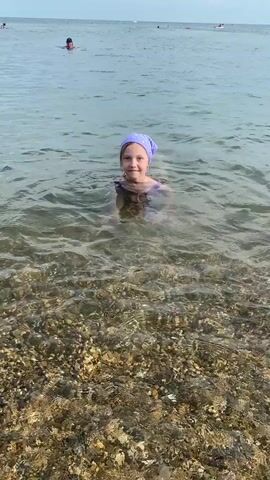 A young girl with a purple bandana and a purple swimsuit is swimming in the ocean. She is smiling and looking at the camera. The water is clear and you can see the pebbles on the seabed. In the background, there are other people swimming and sunbathing. The sky is cloudy, suggesting it is either early morning or late afternoon. The location appears to be a beach in Myskhako, Russia.