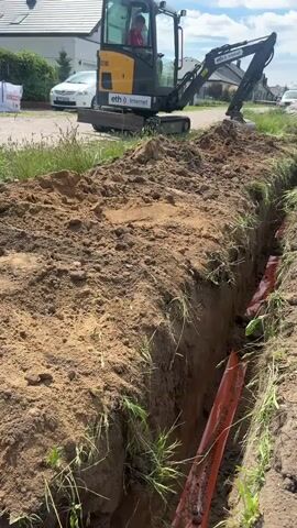 A miniature excavator is digging a trench in the dirt. The excavator is yellow and black, and it has a driver inside wearing a red shirt. The trench is deep and narrow, and there is an orange pipe laid inside it. The environment is a suburban area with houses and a road in the background. The sky is partly cloudy.