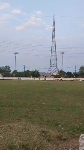 A wide shot shows an outdoor grassy field with several people playing a game, possibly soccer, in the background. A large metal tower dominates the right side of the frame, with power lines extending across the sky. Two lampposts are visible on the field. In the foreground, a dark-colored motorcycle is parked on the grass. The sky is partly cloudy.