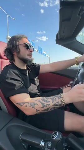 The video shows a man driving a convertible Mercedes-Benz SLK on a highway on a sunny day. The man is wearing sunglasses, a black T-shirt, and black shorts. He has a beard, long hair, and tattoos on his arm. He is wearing a gold watch and a gold bracelet. The car has a red interior. The highway is relatively empty. There are some buildings visible in the distance. The camera is positioned inside the car, providing a driver's perspective.