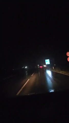 The video captures a first-person perspective from inside a moving vehicle driving on a wet road at night. The view is through the front windshield, looking forward down a dark highway. The road surface is reflective due to being wet, showing reflections of distant lights. A solid white line is visible on the left side of the current lane. In the midground, several vehicles are identifiable by their red taillights and white headlights, scattered in the distance. Faint streetlights line the left side of the road. On the right side of the road, a blue rectangular road sign with white text is clearly visible, followed by a small red pole further down the shoulder. The overall scene is dark, with artificial lights providing the primary illumination.