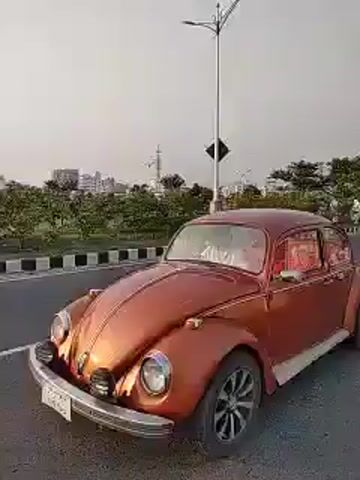 The video shows a red classic Volkswagen Beetle driving down a road. The sun is setting in the background. The Beetle is followed by a group of people walking in white clothing, some carrying bags or backpacks. They are walking on the side of the road alongside the car. The video appears to be taken from another vehicle, possibly following the Beetle. The scene seems to be taking place in an urban environment, with streetlights, trees, and buildings visible. The video continues as the Beetle and the group of people pass under a bridge.