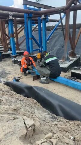 Two construction workers are on a sandy construction site. The worker on the left wears an orange hard hat and an orange high-visibility jacket, crouched down with a tool in hand. The worker on the right wears a gray jumpsuit and a green and white high-visibility stripe on their jacket, also crouched and appearing to be welding or using a grinder. They are working on a blue metal beam that is part of a larger structure of steel beams and pipes overhead. A roll of black material, possibly a liner or membrane, is laid out in the foreground. The environment appears to be an industrial or infrastructure construction area with exposed concrete bases and large metal supports.