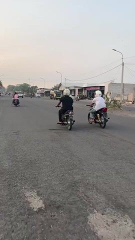 The video shows a paved road with multiple motorcycles and scooters. The sky is overcast. In the foreground, a person is riding a black scooter, with their legs visible. Further down the road, two individuals on motorcycles are moving away from the camera. One is wearing a black shirt and the other a white shirt. Several other motorcycles and scooters are visible in the distance. Buildings and utility poles line the right side of the road. The overall scene depicts traffic on a road in what appears to be a moderately developed area.
