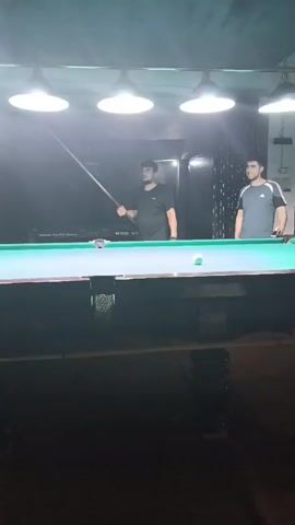 The video shows two young men in a dimly lit room that appears to be a bar or billiard hall. Both are dressed casually. One man on the left, wearing a black t-shirt, is holding a pool cue and appears to be about to take a shot. The second man, wearing a grey t-shirt, stands to the right, observing. A pool table with several balls is visible between them. In the background, a television screen displays a sports game, possibly soccer. The room is decorated with dark curtains.