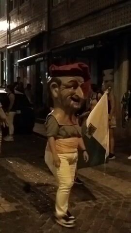 A person in a giant head costume with a red beret, a tan shirt, and yellow pants walks down a cobblestone street at night. The costume has a wide smile and a large nose. The person is holding a white flag with a dark green stripe. Other people are walking behind and around the costumed person, some of whom are holding flags or banners. Buildings with dark facades line the street. The lighting is dim, indicating it is nighttime.