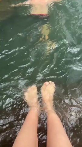 The video shows a person standing with their bare feet in a body of water, presumably a river or lake. The water is clear and slightly agitated. In the distance, a man swims towards a waterfall. The camera pans upwards to focus on the waterfall, which cascades over a rocky surface. The overall scene appears to be a natural, outdoor setting.