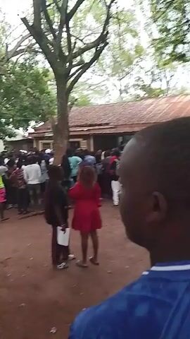 A crowd of people is gathered in an outdoor area with trees. The ground appears to be dirt or reddish soil. There's a low building in the background with a reddish roof. Many people are standing, and there is general movement. Several people are wearing brightly colored clothing, including a woman in a red dress. The camera is moving and appears to be handheld, capturing the scene from within the crowd.