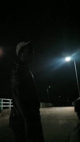 The video is a series of dark, shaky, handheld shots taken at night, likely outdoors. The first shot shows a person wearing a cap and a patterned shirt, silhouetted against a bright street light. The background appears to be a road or bridge with streetlights. Subsequent shots are more obscured, showing glimpses of people and a shirt with text that appears to be "RAHAK" or similar. One shot shows a hand holding a mobile phone, illuminated by its screen. The overall impression is of a casual, possibly social gathering at night in a somewhat urban or suburban environment.