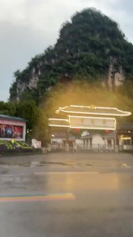 A view of a mountain and an entrance to a Chinese complex on a rainy day. In the background, a mountain covered in green foliage rises. The mountain has exposed rock faces on its sides. In the foreground is an entrance to a complex with a traditional Chinese architectural style. The entrance has a sign with Chinese characters, and it is illuminated with warm lights. A small building is visible behind the entrance. The foreground is a wet road reflecting the lights. Vehicles drive across the road. The sky is overcast and grey.
