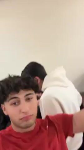 A low-angle video shows two young men indoors. The first man, wearing a red t-shirt and a black watch, has curly brown hair and light skin. He moves around, initially with his hand on his forehead. The second man, wearing a white hoodie, appears later in the video and makes a peace sign with his fingers. The background features a white wall with a window, a black leather couch, and a white pillow. Music plays throughout the video. The second man dances alongside the first man.