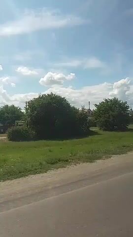 The video shows a rural landscape with a dirt road in the foreground and trees and houses in the background under a partly cloudy blue sky. The perspective suggests the video is being filmed from a moving vehicle, likely on the dirt road, as the scene shifts slightly from left to right. There are no people or specific actions visible, just a static view of the environment. The houses appear to be simple, typical rural dwellings. The trees are lush and green, indicating a temperate climate, likely during the warmer months. The overall impression is one of a peaceful, bucolic setting.