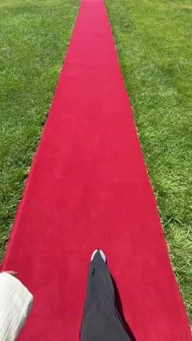 The video shows a person walking down a red carpet laid out on a grassy lawn. The carpet extends towards a large building in the distance, which appears to be a stately home or event venue. The sky is blue with scattered clouds, and trees are visible on either side of the lawn. The camera perspective is from behind the person walking, looking forward along the carpet. The person is wearing dark pants and light-colored shoes. The setting suggests an outdoor event or a special occasion.