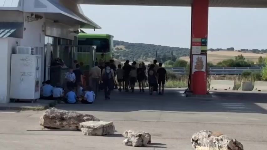 A group of people, mostly young, are gathered at a gas station. Some are sitting on the ground near a building, while others stand in small groups or walk around. A green bus is parked near the building. A red gas pump is visible to the right. The scene appears to be outdoors, with a hilly, rural landscape in the background. The weather seems to be sunny.