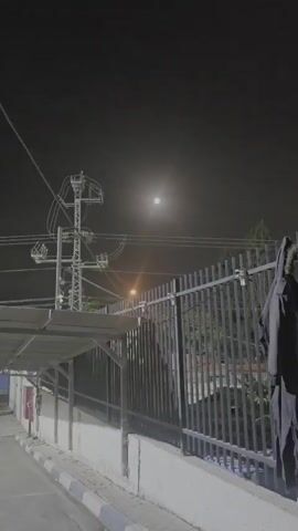A low-angle shot captures a nighttime scene. A bright full moon is visible in the hazy dark sky. Below the moon, three parallel power lines stretch across the frame. To the left, a utility pole with numerous insulators and wires stands tall. Further left, a covered walkway with a slanted roof is partially visible, and a red fire extinguisher is mounted on a wall beneath it. In the center of the frame, a street lamp emits a warm orange glow. A black metal fence with vertical bars runs along the bottom of the image. A dark garment hangs on the right side of the fence. A paved area with a white and grey curb is in the foreground.