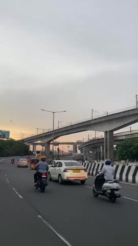 The video shows a highway scene with an elevated train track overhead. Several vehicles, including cars and motorcycles, are traveling on the highway. There is a traffic sign with directions to Koyambedu and Poonamallee. The sky is overcast, and the lighting suggests it is either early morning or late afternoon. The overall atmosphere is that of a busy urban commute.