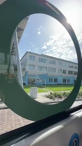 The video shows a view of a building through a circular green frame. The building has three floors and is painted white with light blue accents. A green lawn is in front of the building. The word "WoonArk" is visible on the front of the building. A dark-colored car is parked in front of the building and then drives off. A blue sky with scattered clouds is in the background, with the sun shining. The video is shot from inside a moving vehicle, likely a bus or tram, as evidenced by the interior features of the vehicle visible at the bottom of the frame. The road in front of the building is paved with bricks.